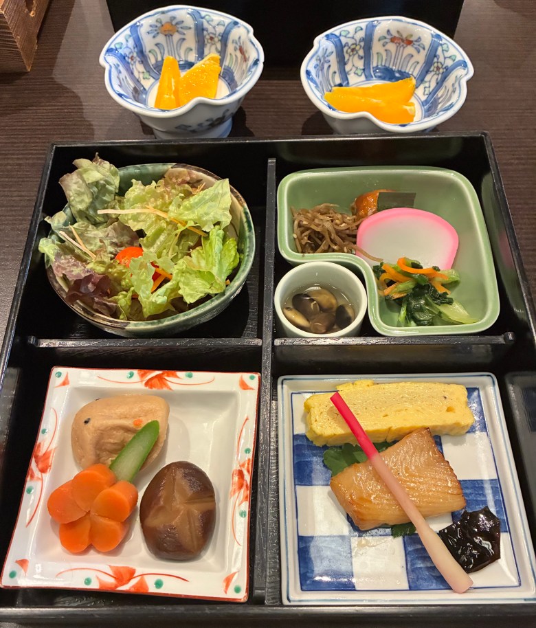 photo of a tray of food with four boxes holding a salad, pickled vegetables, orange and brown blobs, and a small egg omelette slice next to a light brown piece of white fish