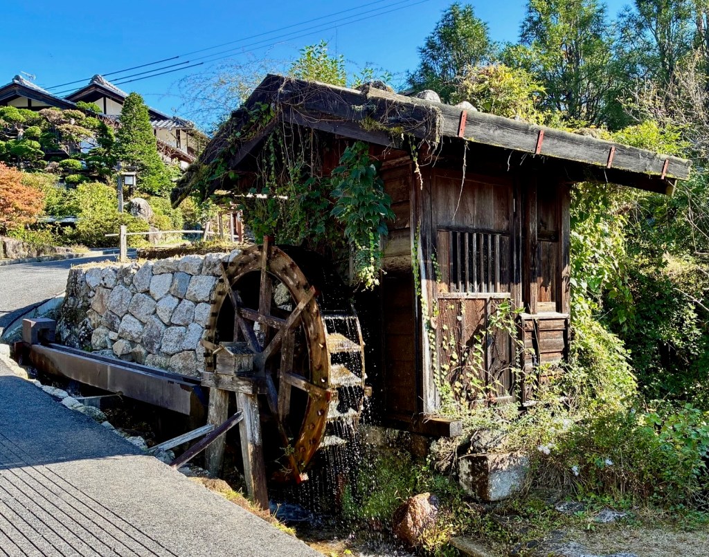 photo of a dark brown wooden water wheel spinning with water dripping off, next to a cobblestone road with blue sky above