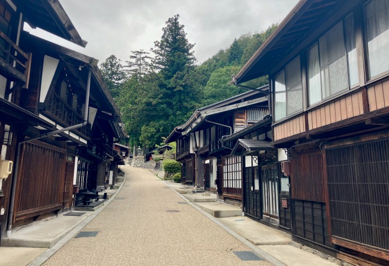 photo of a sandy colored street curving up a hill with green trees at the end, lined by dark brown wooden two story buildings with white windows