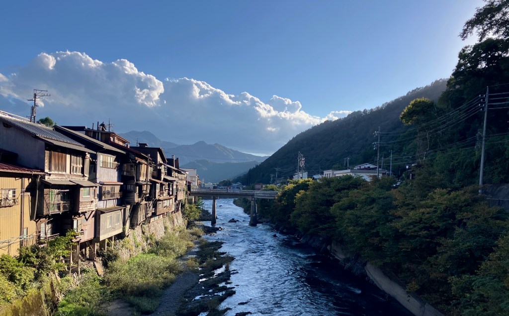 photo of a river running next to a row of three-story brown buildings on stilts, with a small bridge across it and mountains and clouds in the distance