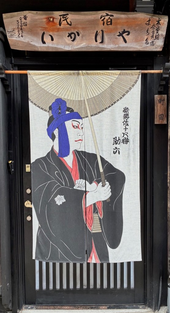 photo of a doorway with a wooden Japanese sign across the top and a banner painting of a samurai holding an umbrella wearing a blue bandana and red and black kimono