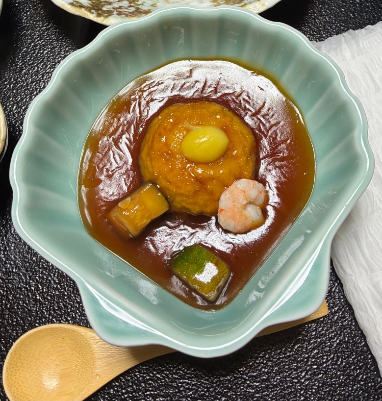 close-up photo of a light green, scallop-shaped dish holding a glistening, gelatinous liquid with blobs of orange, green and pink in it.