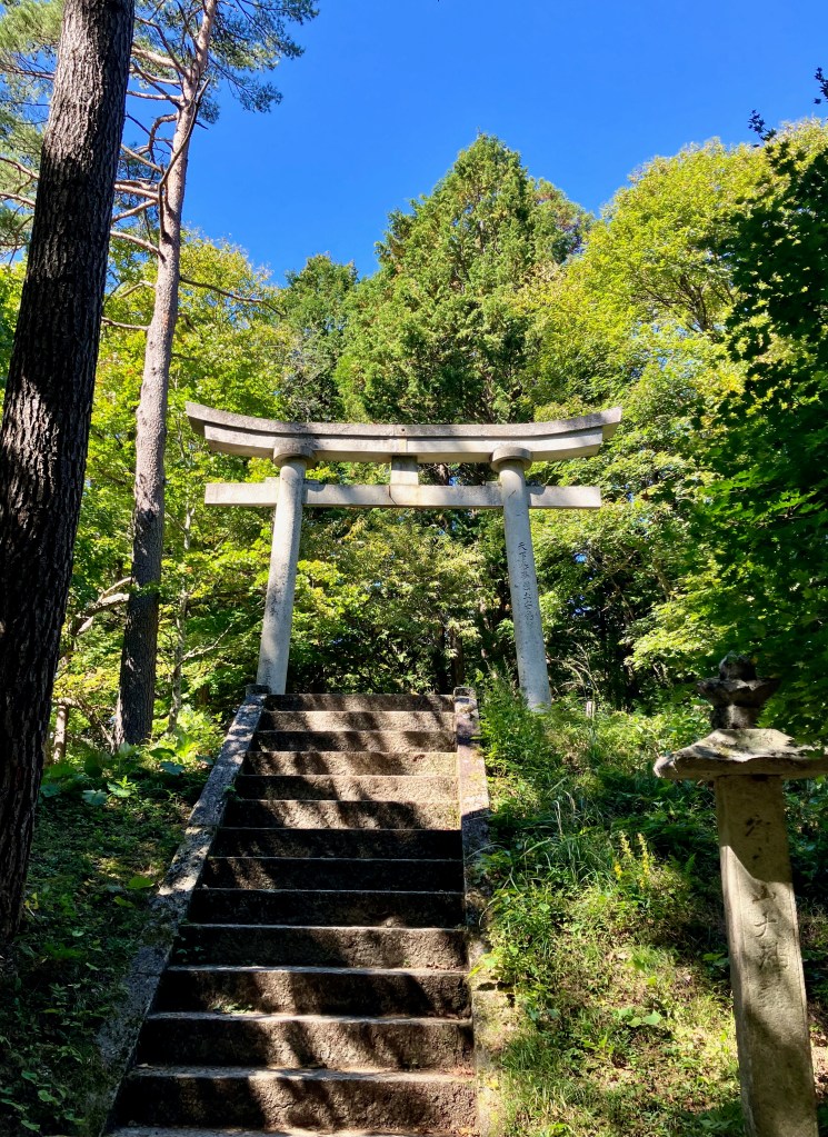 photo of a white stone Japanese arch at the top of stone steps with green trees all around and blue sky behind